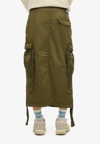 Olijfgroen cargorok met meerdere zakken en hangende banden, gedragen met blauw en wit gestreepte sokken en beige sneakers.