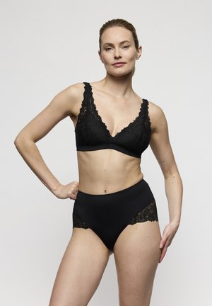 Vrouw die een zwart kanten beha en hooggetailleerd ondergoed draagt, staand met één hand op haar heup en een neutrale uitdrukking tegen een witte achtergrond.