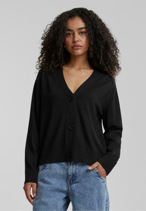 Svart cardigan med V-hals, frontknapper og ribbede mansjetter. Brukt over lyseblå jeans, har en avslappet passform og myk tekstur.