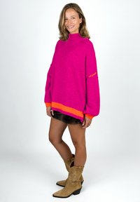 Frau steht lächelnd da, trägt einen hellrosa übergroßen Pullover mit orangefarbenen Akzenten, einen schwarzen Rock und hellbraune Cowboystiefel vor einem weißen Hintergrund.