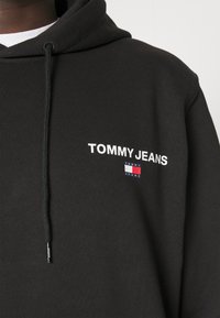 Felpa nera con cappuccio realizzata in tessuto testurizzato e dotata di tasca frontale. Presenta il logo "TOMMY JEANS" ricamato in bianco e un piccolo dettaglio a forma di bandiera colorata.