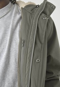 Olivgrüne Jacke mit weich beigegefüttertem Fleece; enthält einen dicken Reißverschluss und Druckknöpfe; getragen über einem hellgrauen, kurzärmeligen Hemd.