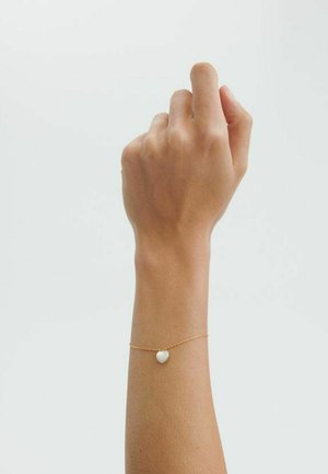 Rechter Arm erhoben, Finger leicht gekrümmt, trägt ein filigranes goldenes Armband mit einem kleinen weißen Herzanhänger am Handgelenk.