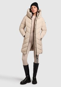 Beige puffjakke med et chevronmønster, stor hætte og lynlåse. Kombination med et tætsiddende beige sæt og knæhøje sorte støvler.