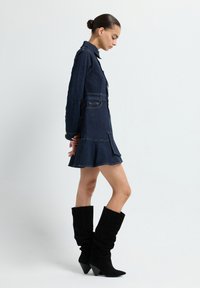 Robe en denim foncé avec une jupe évasée et un devant boutonné, associée à des bottes noires en daim montant jusqu'aux genoux avec un bout pointu et un talon cône.