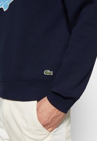 Personne portant un sweat bleu marine avec un petit logo crocodile vert près de l’ourlet, main reposant dans la poche d’un pantalon blanc cassé.