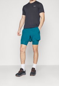 Mies, joka käyttää mustaa lyhythihaista urheilupaitaa, turkooseja kerroksittain leikattuja shortseja, valkoisia urheilusukkia ja mustia juoksukenkiä, seisoo tasaisella taustalla.