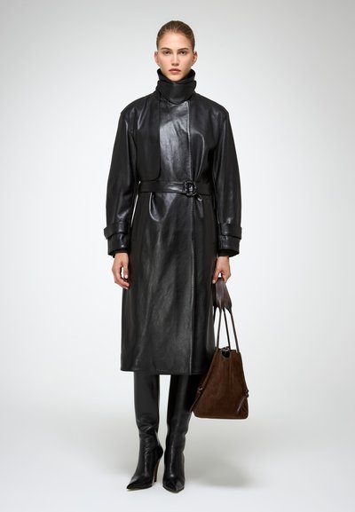 Donna che indossa un cappotto di pelle nero con cintura, stivali neri con tacco alto e tiene in mano una borsa in suede marrone su uno sfondo neutro.