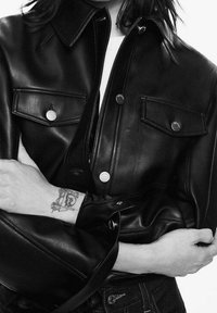 Czarna skórzana kurtka z kołnierzem, zapięciem na guziki i dwiema przednimi kieszeniami. Ramiona modela są skrzyżowane, a na nadgarstku widać tatuaż.