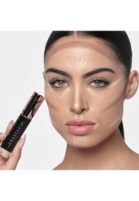 Zwart- en roségouden verpakking van het highlighterproduct van Anastasia Beverly Hills, vastgehouden naast het gezicht van een model met aangebrachte contourlijnen in verschillende tinten.