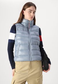 Gilet rembourré bleu clair avec col haut, fermeture éclair sur le devant et poches latérales ; comporte un logo rond sur la poitrine et une texture lisse.