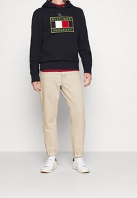 Felpa con cappuccio blu navy decorata con logo, tasca a marsupio e cappuccio con lacci. Abbinata a pantaloni beige e sneakers bianchi. Look moderno e casual.