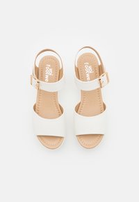 White faux leather sandals with two wide straps, a mid-height heel, and a buckle detail. Soft beige insole with stitching accents.