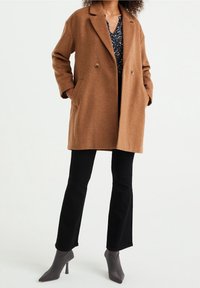 Brązowy płaszcz z mieszanki wełny z wyciętym kołnierzem, zapięciem na dwa guziki, bocznymi kieszeniami i luźnym krojem; zestawiony z czarnymi spodniami dzwonami i szarymi szpilkami.