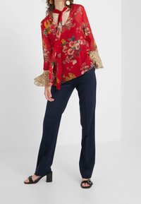 Blusa roja con flores y cuello atado, puños con estampado de leopardo; combinada con pantalones navy a medida y sandalias de tacón negras.