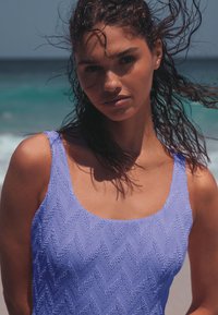 Donna con capelli scuri bagnati e scompigliati dal vento che indossa un costume da bagno in pervinca texturizzato, in piedi vicino all'oceano in una giornata di sole.