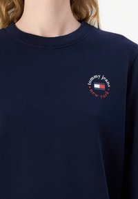 Person wearing navy blue sweatshirt with “tommy jeans new york” text and red, white, and blue rectangular logo on chest.