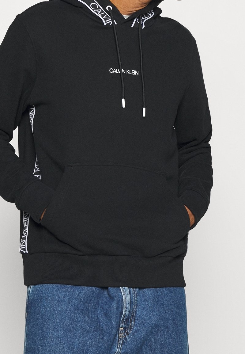 Homme portant un sweat à capuche noir Calvin Klein avec logo sur la poitrine, bordure de la capuche et bandes latérales, associé à un jean bleu, mains dans la poche avant.