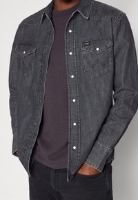 Harmaa farkkupaita, jossa on kaksi rintataskua, nepparit ja hieman teksturoitu kangas. Käytetään tumman t-paidan päällä.