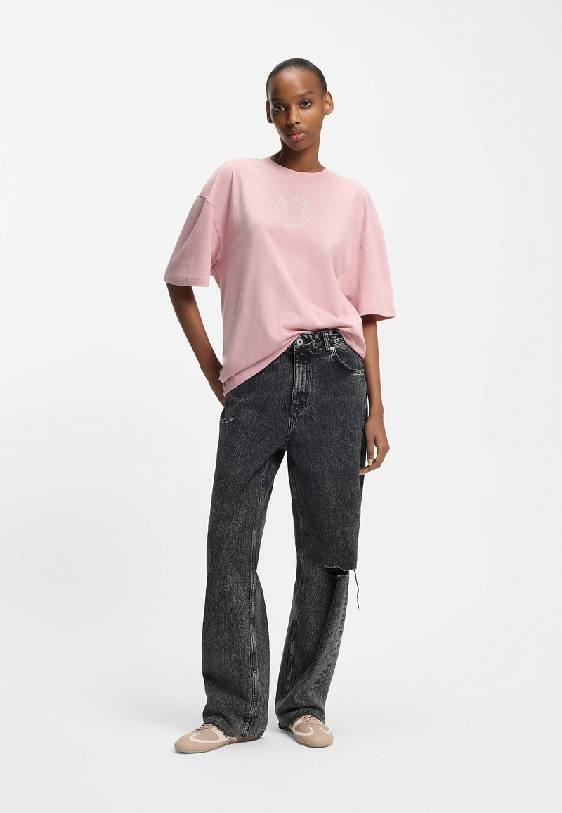 T-shirt rose pâle oversize avec un graphisme circulaire, associée à un jean noir délavé à jambes larges avec des détails usés et des chaussures marron clair.