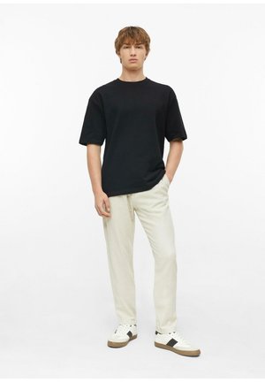 Young man standing in casual outfit with black short-sleeve shirt, light beige pants, and white sneakers with black stripes on white background.