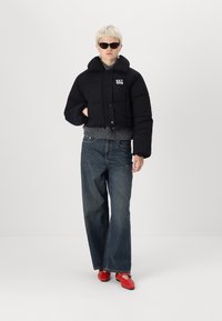 Forkortet sort dunjakke med høj krave, udstyret med en frontlukning med knapper og logo-detalje. Båret over en grå sweater og blå jeans.