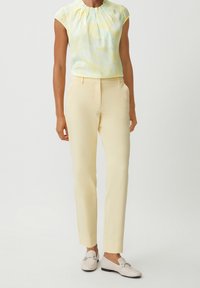 Femme portant un pantalon sur mesure jaune pâle, un chemisier à motifs jaunes clairs et des mocassins crème avec des détails en métal.