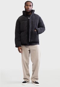 Chaqueta de abrigo de pana negra con cuello alto, que presenta bolsillos frontales y cierre a presión. Llevada sobre una camiseta negra y pantalones beiges.