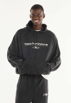 Junger Mann lächelt und steht mit den Händen in den Taschen, trägt einen schwarzen New Balance Hoodie und passende schwarze Jogginghosen.