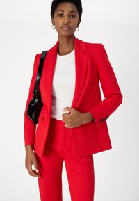 Rød skreddersydd blazer med matchende bukser, som har en glatt tekstur og strukturert design. I kombinasjon med en hvit ribbestrikket topp og en svart lærveske.