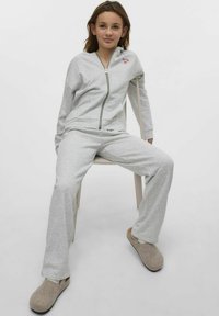 Grå zip-up hoodie och matchande byxor i mjukt material, med en liten röd logotyp. Modellen bär beige inneskor och sitter på en stol.