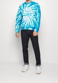 Sudadera azul con efecto tie-dye y logo blanco, combinada con jeans oscuros y zapatillas blancas. Presenta mangas largas y puños de canalé.