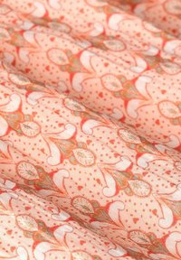 Leichtes Pfirsichfarbendes Gewebe mit einem floralen Muster, das komplizierte Spiralen und Blätter in Koralle und Grün zeigt, vor einem gepunkteten Hintergrund.