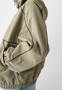 Osoba w dużej beżowej kurtce z czarnymi wykończeniami, elastycznymi mankietami oraz jasnoniebieskich dżinsach, stojąca na tle jednolitym.