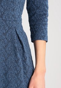 Robe bleue en tissu texturé avec un motif floral en relief, une coupe ajustée et des manches trois-quarts. Finitions détaillées avec des coutures.