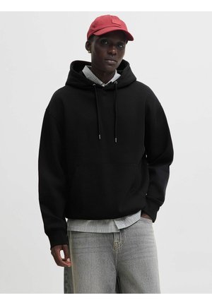 Jong persoon met een zwarte hoodie, grijs geruit overhemd, lichtgrijze spijkerbroek, rode pet en gouden oorbel, staand tegen een effen achtergrond.