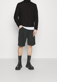 Sort syntetisk zip-up jakke med høj krave, kombineret med mørkegrå plisserede shorts og sorte læder ankelstøvler.