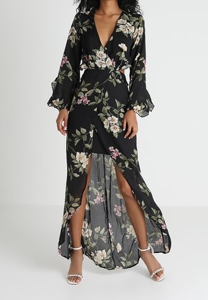 Femme portant une robe portefeuille noire à motifs floraux, à manches longues et avec de hautes fentes à l'avant, associée à des talons ouverts blancs.