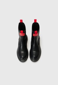 Botas de tornozelo em pele preta com uma textura lisa, apresentando puxadores vermelhos e marcas laterais, além de uma sola espessa e texturizada.