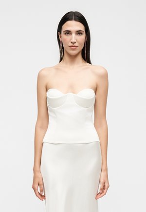 Femme aux longs cheveux foncés portant une longue robe blanche bustier et des boucles d'oreilles pendantes, debout devant un fond clair uni.