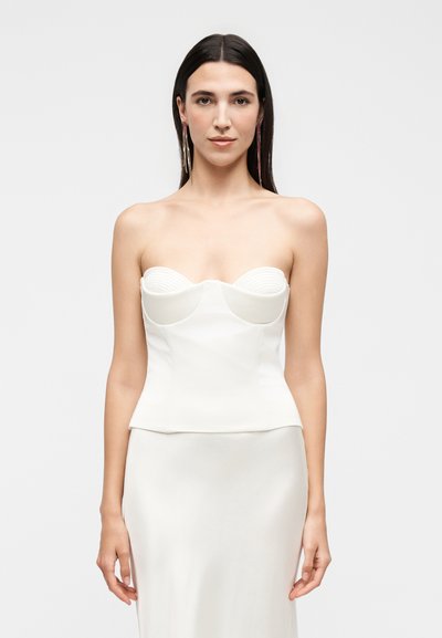 Femme aux longs cheveux foncés portant une longue robe blanche bustier et des boucles d'oreilles pendantes, debout devant un fond clair uni.