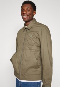 Alyvų žalia užtrauktuku užsegama striukė, pagaminta iš lygios medžiagos, su apykakle, šoninėmis kišenėmis ir mažuoju žaliu logotipu krūtinėje.