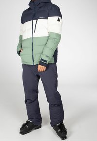 Padded jacket with navy, white, and green color blocks. Features a zip closure, hood, and elastic cuffs. Worn with dark pants and ski boots.