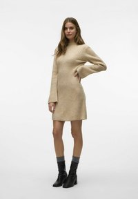 Robe en tricot beige avec une texture côtelée, des manches longues évasées, un col haut et un ourlet légèrement évasé, associée à des bottines noires et des chaussettes grises.