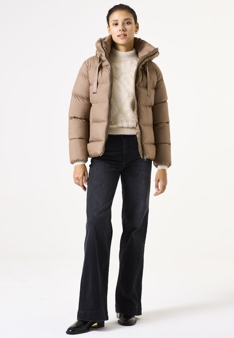 Chaqueta de plumas marrón con capucha, con gruesas costuras horizontales, dos bolsillos frontales, combinada con un suéter crema y jeans negros de campana.