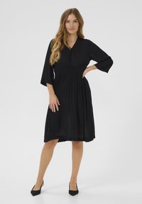 Robe noire avec un décolleté en V, des manches trois-quarts et une taille smockée. Tissu fluide qui tombe jusqu'au mollet, texturé avec un subtil éclat.
