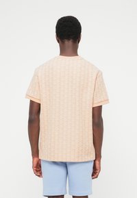 Vyras dėvintis šviesiai persikinės spalvos trumpomis rankovėmis margintą marškinių palaidinę ir šviesiai mėlynas šortas, stovintis nugarą atsukęs priešais paprastą baltą foną.