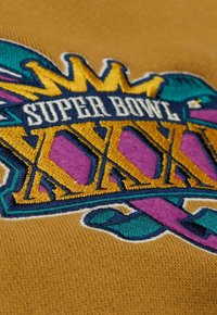 Logo ricamato del Super Bowl XXXIX in blu, viola e oro su un tessuto giallo senape testurizzato; il design presenta una scritta audace e accenti a forma di corona.