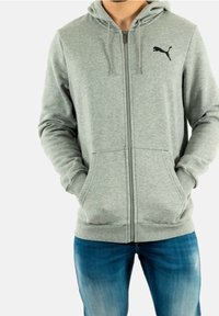 Gray zip-up hoodie in soft textile, featuring a kangaroo pocket, drawstring hood, and black brand logo on the chest. Paired with blue jeans.