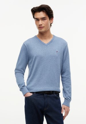 Νέος άνδρας που φοράει ανοιχτό μπλε πουλόβερ με σχήμα V και σκούρα μπλε τζιν, στέκεται με το ένα χέρι στην τσέπη του μπροστά σε λευκό φόντο.